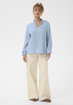 Top de manga larga azul claro con un collar con volantes, combinado con pantalones de pierna ancha crema. Textura suave con un corte relajado y detalles de cordón.