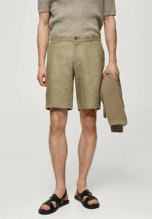 Mand iført beige shorts og brun strikket top, der holder en matchende sweater, med sorte sandaler på en ensfarvet baggrund.