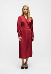 Robe rouge en satin avec des manches longues, un col à volants et une taille à nouer. Texture lisse ; longueur genou avec une silhouette droite et fluide.