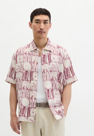 Mężczyzna w koszuli z krótkim rękawem na guziki z różowo-białymi kwiatowymi i paskowanymi wzorami, beżowych spodniach i brązowym pasku.