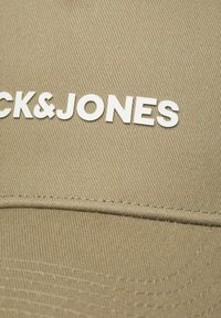 Beżowa czapka z widoczną teksturowaną tkaniną, z białym logo "BLACK&JONES" na przodzie i subtelnym przeszyciem przy krawędziach.