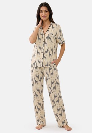 Frau steht barfuß, trägt beige Knopf-Pyjamas mit schwarzen und weißen Giraffen gemustert, eine Hand in der Tasche und lächelt.