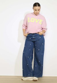 Rozā adīta džemperis ar uzrakstu "Mīlestība" dzeltenā krāsā, savienots ar platām denima biksēm ar ziedu izšuvumiem. Izskatu papildina neitrālu toņu sporta apavi.