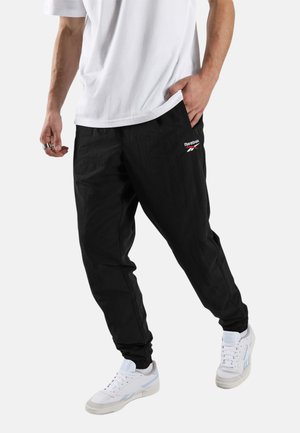 Homme portant un jogging noir Reebok, des baskets blanches et un t-shirt blanc, une main dans la poche et l'autre détendue le long du corps.