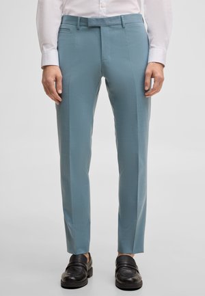 Mężczyzna ubrany w jasnoniebieskie, szyte na miarę spodnie, białą koszulę i czarne skórzane mokasyny, stojący na jednolitym tle.