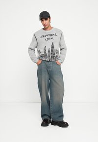 Grijze trui met zwart stadslandschap, gecombineerd met lichtblauwe wijde jeans. Model draagt ook een zwarte pet en robuuste zwarte schoenen.