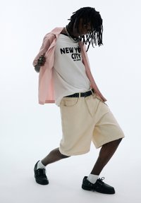 Camicia rosa a maniche corte con bottoni sopra una maglietta bianca con stampa nera, pantaloni beige larghi, scarpe nere e calze bianche. Dettaglio a catena nella cintura.