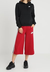 Svart huvtröja med framficka och vit Nike-logotyp; röda vida sweatpants med sidostrippar och Nike-logotyp; svarta sneakers.