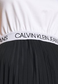 Gros plan d'une ceinture blanche avec un texte noir "Calvin Klein Jeans" sur une jupe plissée noire ou un pantalon.