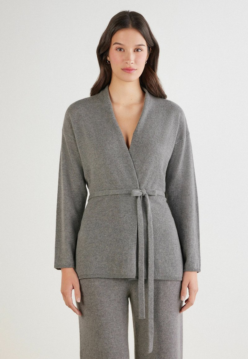 Cárdigan envolvente gris de punto, hecho de material suave, con un cinturón en la cintura, mangas largas y un diseño de escote en V.