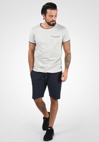 T-shirt gris clair à manches courtes avec une poche et des bordures foncées, associé à un short bleu marine foncé et des baskets de sport noires, se tenant sur un fond dégradé.