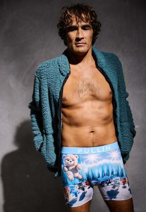 Boxers à imprimé ours en peluche bleus avec une taille bleu clair étiquetée "PULLIN", portés sous une veste zippée verte et duveteuse.