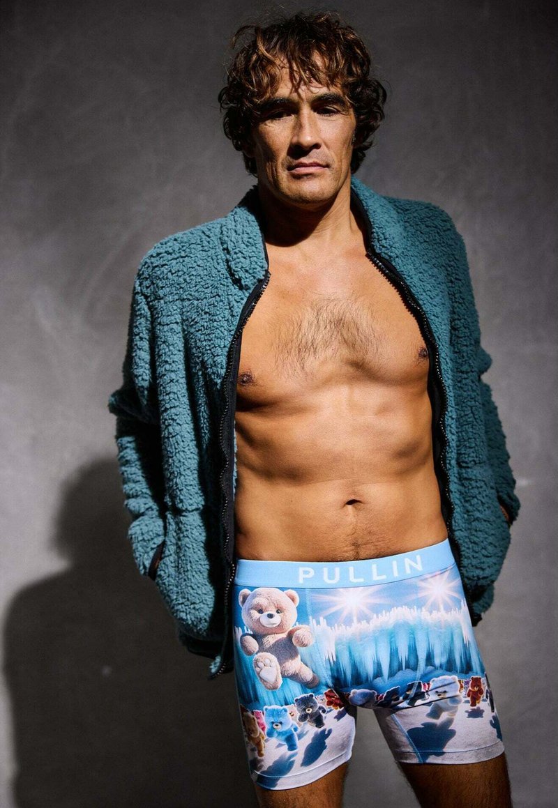 Boxers à imprimé ours en peluche bleus avec une taille bleu clair étiquetée "PULLIN", portés sous une veste zippée verte et duveteuse.