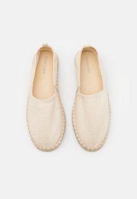Zapatos sin cordones de lona beige con una superficie texturizada, punta redonda y suelas trenzadas de yute. Diseño sencillo sin herrajes visibles.
