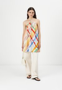 Robe halter colorée avec des rayures diagonales en jaune, rouge, bleu et vert ; associée à un pantalon large blanc et des tongs noires.