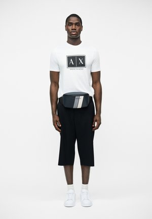 Mand iført hvid Armani Exchange t-shirt, sorte knælange shorts, hvide sneakers og en sort hoftepose med hvide striber.