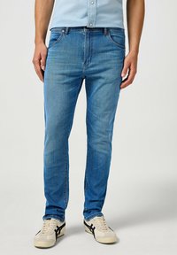 Jeans de mezclilla azul claro con corte slim, que cuentan con bolsillos frontales y cierre de cremallera. Llevados con zapatillas beige y una camisa azul clara.