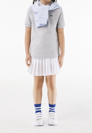 Camisa polo gris de manga corta, suéter azul claro sobre los hombros, falda plisada blanca, calcetines a rayas azules y blancas, zapatillas blancas.