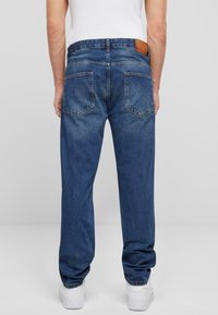 Blugi din denim albastru cu croi drept, două buzunare la spate și o etichetă din piele maro pe talie. Adidași albi vizibili la picioare.