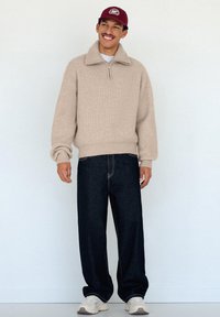 Maglione beige lavorato a maglia con collo a zip, abbinato a jeans in denim scuro e larghi. Scarpe da ginnastica bianche e cappellino da baseball rosso completano l'outfit.