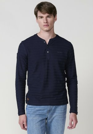 Tumesinine pika varrukaga henley särk, millel on õrn triibuline tekstuur, kolme nupuga sõlm ja logo detail rinnal. Kanda helesiniste teksadega.