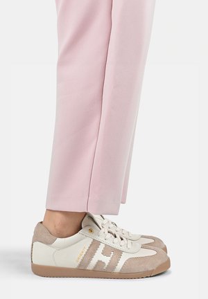 Persoon die witte en beige sneakers draagt met "H" logo, gecombineerd met lichtroze broek, staand op een witte ondergrond.