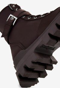 Smeđe gležnjače s gornjim dijelom od glatke kože, sadrže srebrnu kopču i detalje vezanja, te debeli đon s profilom i brendiranjem.