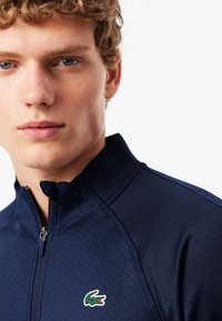 Chaqueta navy con cremallera y cuello alto, fabricada con un tejido texturizado, y un pequeño logo de cocodrilo en verde y rojo en el lado izquierdo.