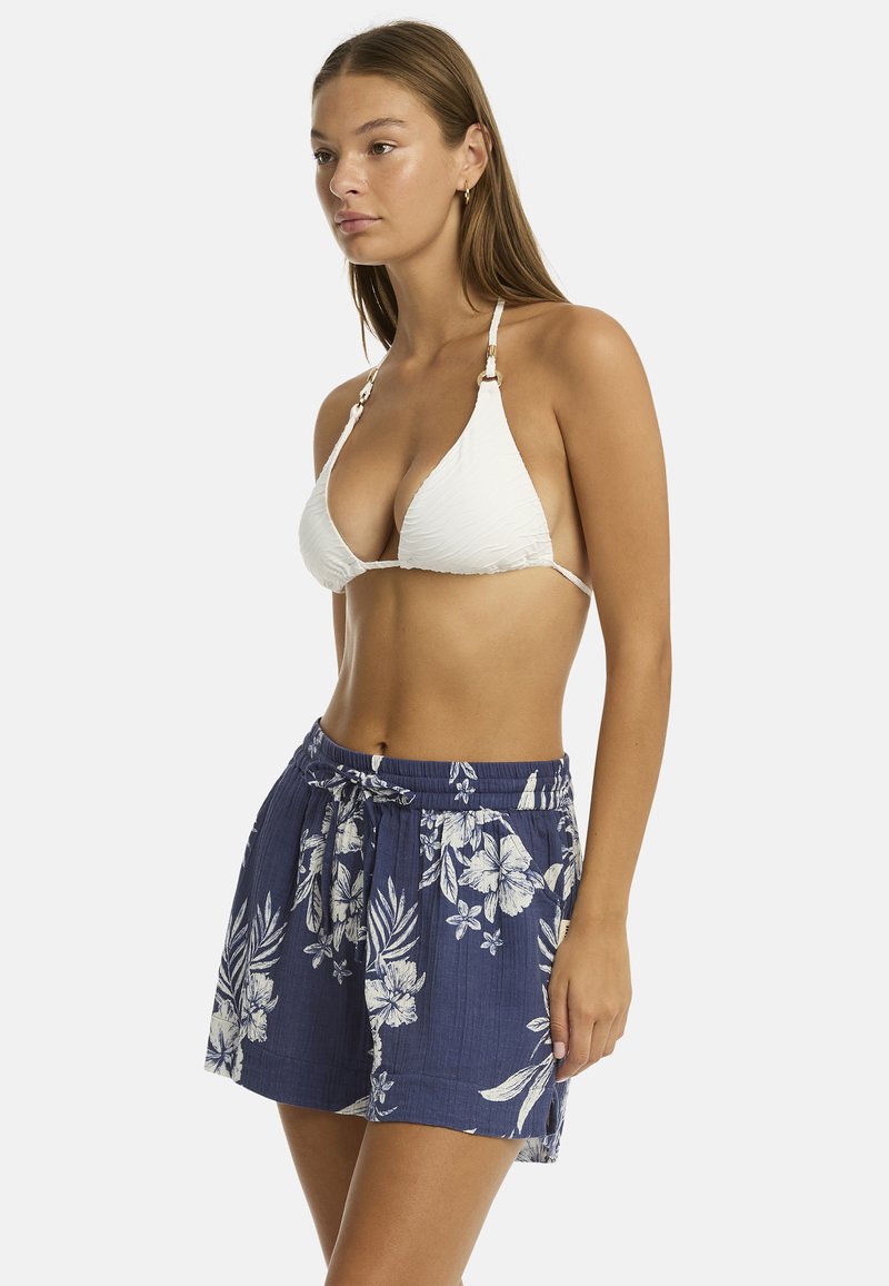 Kobieta z długimi włosami pozuje w profilu, ubrana w biały top bikini i szorty w kwiatowy wzór, prezentując strój gotowy na plażę.