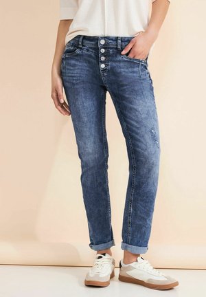 Kobieta w podwiniętych niebieskich jeansach z zapięciem na guziki i białych sneakersach, ręka w kieszeni, stojąca na beżowym tle.