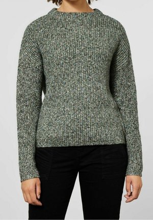 Persoon draagt een groen-grijs geribd gebreide trui met lange mouwen en zwarte broek, kort gesneden bij de nek en heupen.