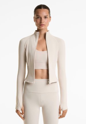 Light beige zip-up jacket with long sleeves. Made of soft material, it features a fitted design and smooth texture, complemented by a high collar.