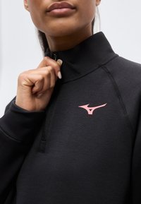 Femme fermant la fermeture éclair d'un pullover de sport noir avec un logo Mizuno rose sur la poitrine, vue en gros plan.