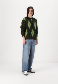 Σκούρο πράσινο πουλόβερ από μαλλί με διαμαντοειδές σχέδιο σε πράσινο και ανοιχτό πράσινο. Συνδυασμένο με ανοιχτό μπλε παντελόνι σε φαρδιά γραμμή και λευκά αθλητικά παπούτσια.