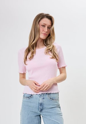 Kvinde med langt blond hår iført en lyserød ribbet kortærmet top og lyseblå jeans med bølgekantede lomme-detaljer mod en ensfarvet baggrund.