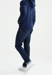 Felpa con cappuccio blu navy con orlo a costine, abbinata a jeans blu scuro e scarpe da ginnastica bianche. La felpa presenta un logo sulla manica e un cappuccio con coulisse.