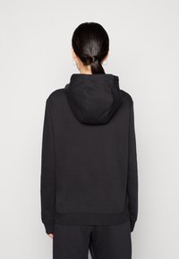 Sudadera negra con bolsillo tipo canguro, tejido texturizado, corte oversize y capucha con cordón. Vista desde la parte trasera, combinada con pantalones oscuros a juego.