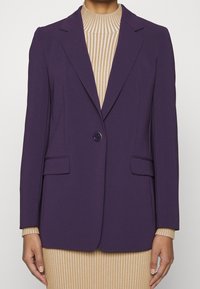 Fioletowa dopasowana marynarka na jeden guzik, z dwoma przednimi kieszeniami i wyraźnie zarysowanymi ramionami, noszona na beżowym prążkowanym topie.