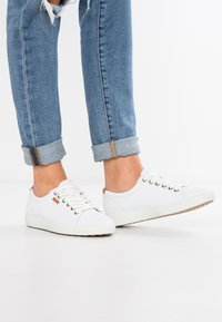 Hvit lerrets sneakers med rund tå, flat såle og beige detaljer. Skolisser og metalløye synlige; sammen med rullet denimjeans.