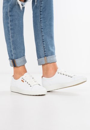 Person iført opbukte blå jeans og hvide lave sneakers med snørebånd, der står på en hvid overflade.