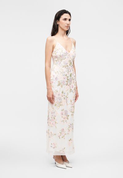 Γυναίκα που φοράει λευκό αμάνικο φόρεμα μακριά έως το πάτωμα με παστέλ κεντημένα λουλούδια και λευκές μυτερές γόβες σε απλό ανοιχτό φόντο.