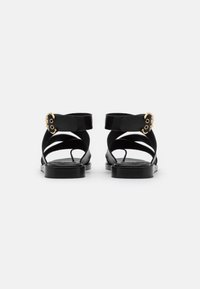 Černé sandály na tenkých páscích mají plochou podrážku, zakřivené kotníkové pásky a zlaté přezky. Hladký kožený povrch s minimálním designem.