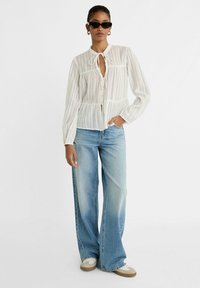 Vit, genomskinlig blus med ränder, knytkrage, knappstängning och långa ärmar, ihopparad med ljusblå utsvängda jeans och sneakers.