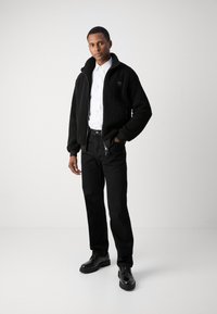 Giacca bomber in pile nera con colletto alto, chiusura con zip e tasche laterali, indossata sopra una camicia bianca e abbinata a jeans neri e scarpe.