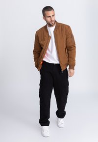 Bruine bomberjack met ritssluitingen, gedragen over een wit T-shirt, gecombineerd met zwarte cargobroek en witte sneakers, tegen een neutrale achtergrond.