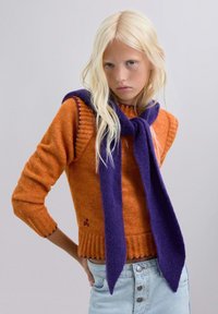 Suéter naranja de punto con dobladillos acanalados, combinado con una larga bufanda morada. Cabello rubio, ojos azules y jeans de mezclilla claros.