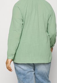 Maglia verde chiaro a maniche lunghe in tessuto texture, con una vestibilità rilassata e polsini con bottoni; abbinata a pantaloni blu chiaro.