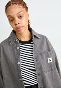 Camicia in denim grigia con chiusura a bottoni e taschino sul petto, indossata sopra una t-shirt a righe bianco e nere. Collo tondo, vestibilità casual.
