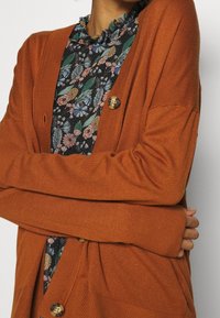 Cardigan color ruggine con maniche lunghe, lavorazione a maglia testurizzata, chiusura frontale con bottoni e vestibilità comoda. Indossato sopra una camicetta con motivo floreale.