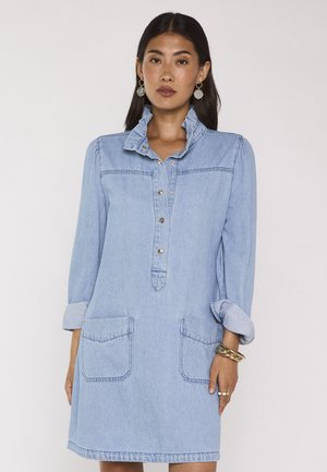 Femme portant une robe en denim bleu clair avec col à volants, manches retroussées, poches avant et patte de boutonnage, debout devant un fond uni.
